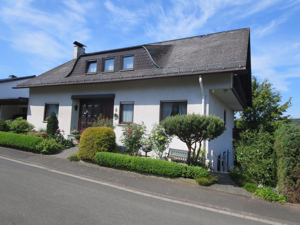 A white cottage-style house with a steep tiled roof, dormer window, and wooden front door, viewed from a quiet road, with landscaped hedges and flower beds along the front and rolling green hills in the distance.